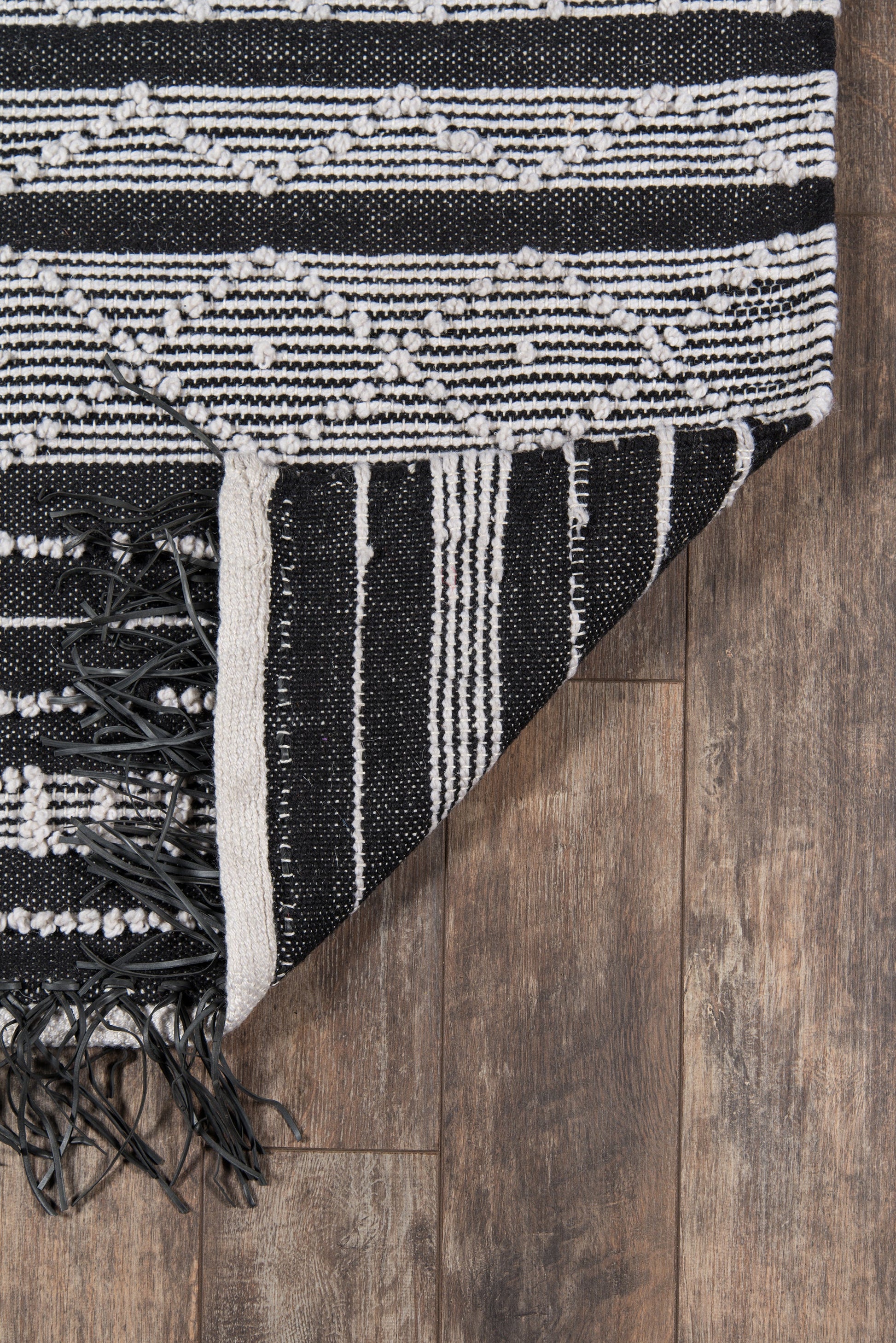 Woven black & white geometric rug with fringe tassels, 5' x 8', folded corner on dark wood floor.
