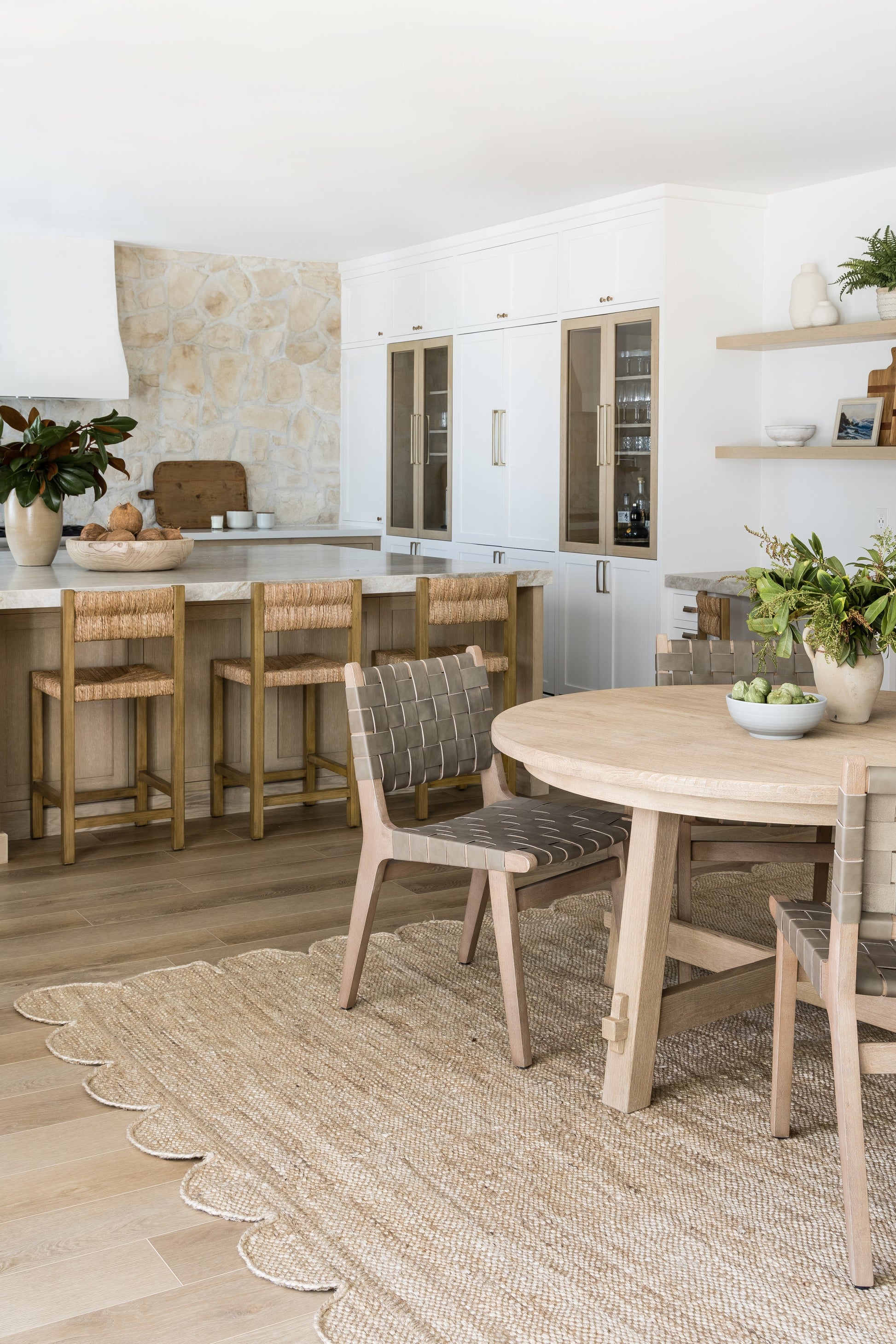 Bright kitchen with white cabinets, natural wood island and stools, stone wall, round wooden dining table on a woven rug.