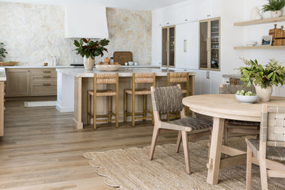 Bright natural-toned kitchen with white cabinets, island and wooden stools, round dining table, woven rug, and potted plants.