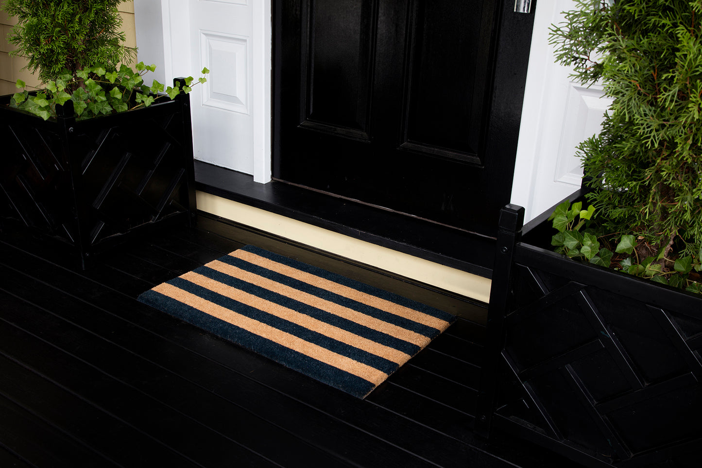 Navy and tan striped doormat on a black wooden porch in front of a black door with white trim, greenery on both sides.