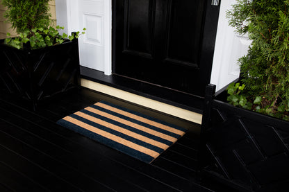 Navy and tan striped doormat on a black wooden porch in front of a black door with white trim, greenery on both sides.