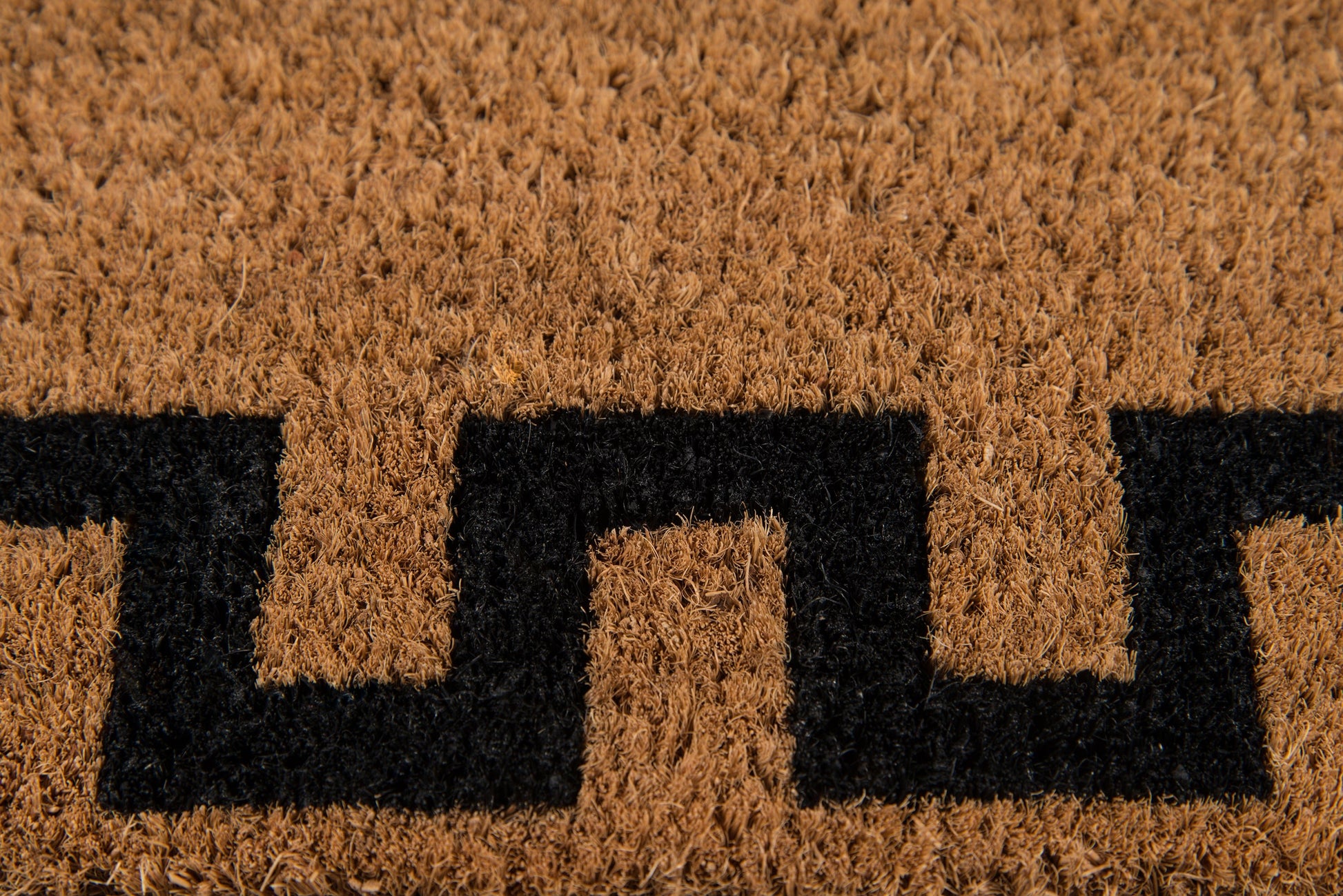 Natural brown coir rug with a black Greek-key geometric pattern; coarse fibrous texture for entrance use.