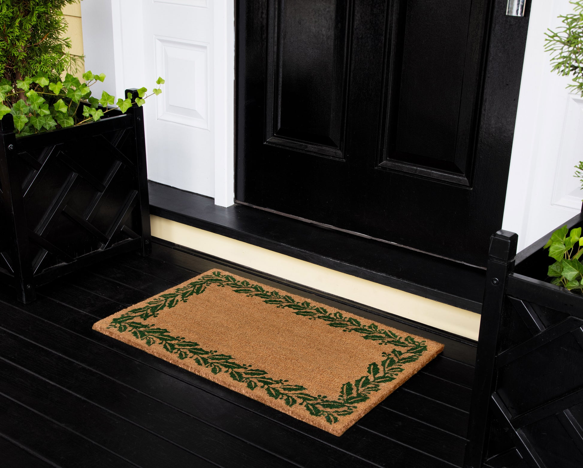 Beige natural-fiber doormat with a green leafy border on a black porch in front of a black entry door.