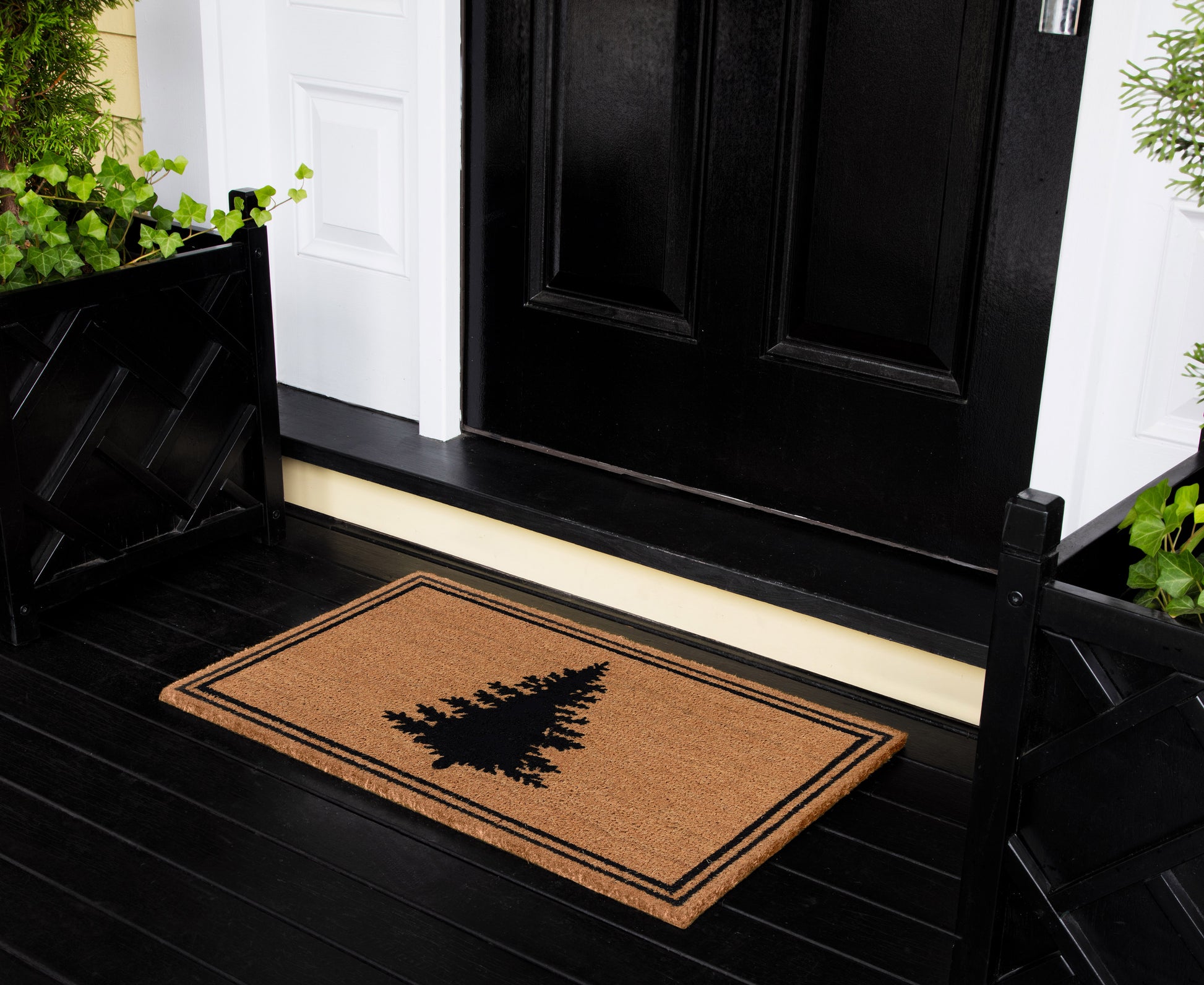 Brown coir front doormat with black pine tree silhouette and double border on a black porch by a white-trimmed doorway.