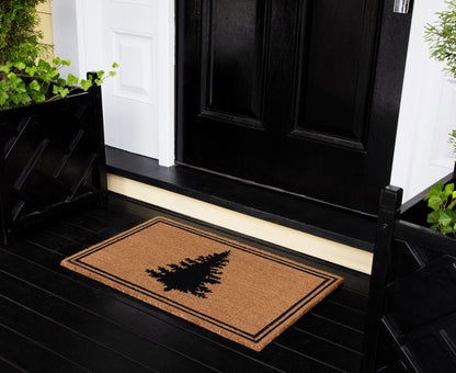 Brown coir front doormat with black pine tree silhouette and double border on a black porch by a white-trimmed doorway.