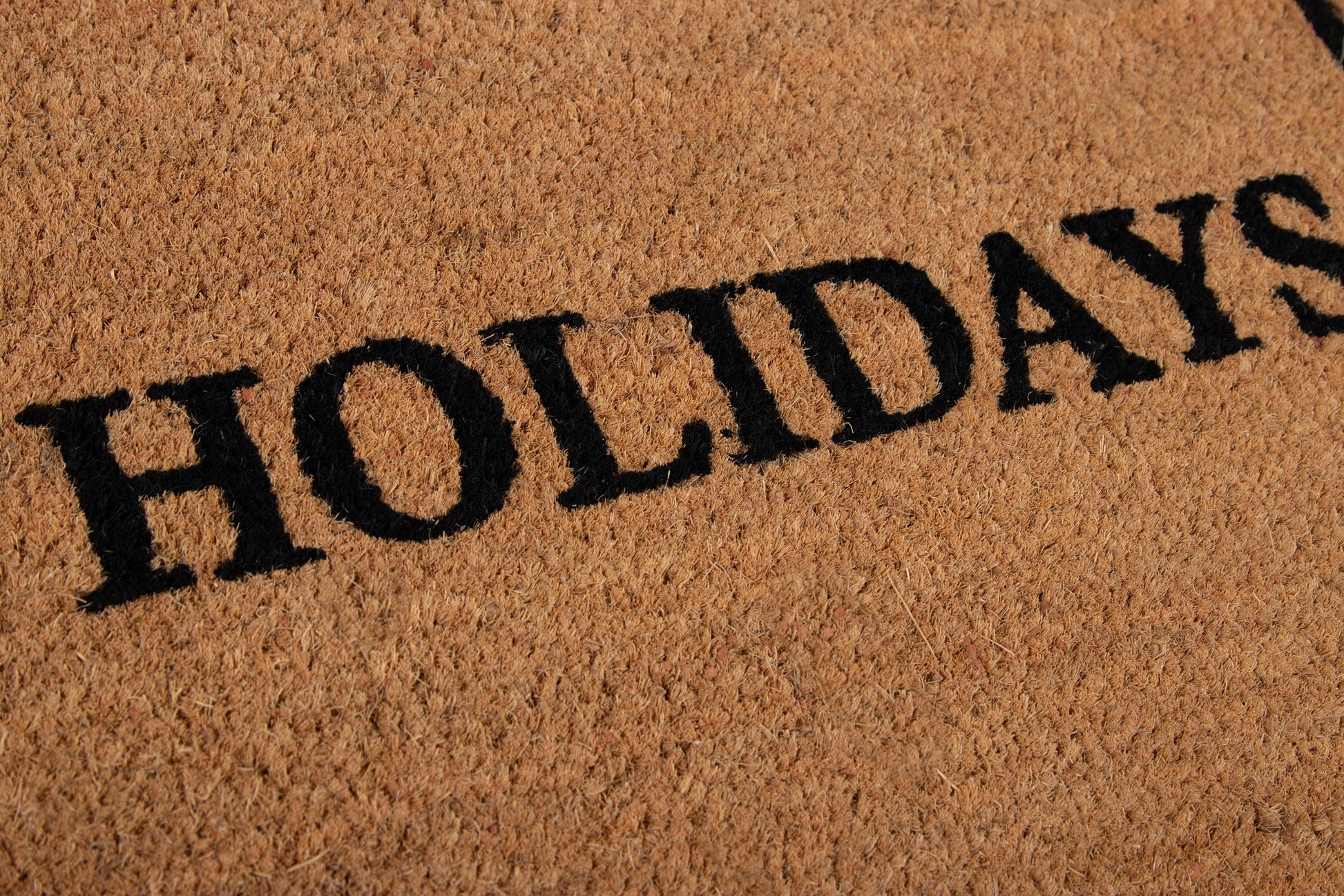 Coir doormat in tan with bold black HOLIDAYS lettering; textured fiber surface.