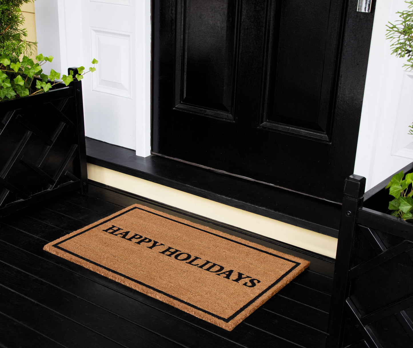 Brown rectangular coir doormat with 'HAPPY HOLIDAYS' in black, at black front door with greenery on the porch.