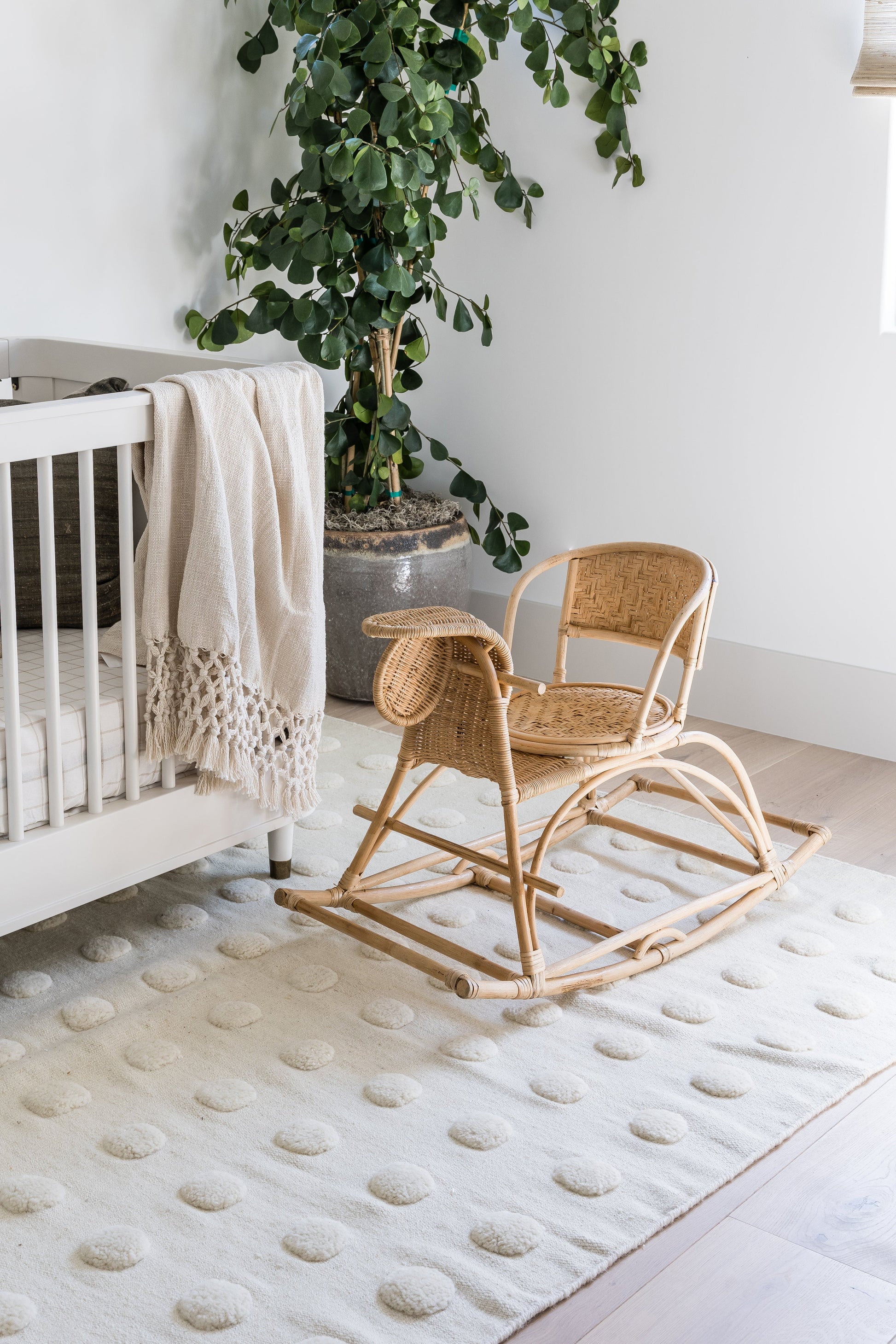 Natural rattan baby rocker on a cream dotted rug beside a white crib with a beige blanket and a leafy potted plant.
