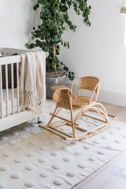 Natural rattan baby rocker on a cream dotted rug beside a white crib with a beige blanket and a leafy potted plant.