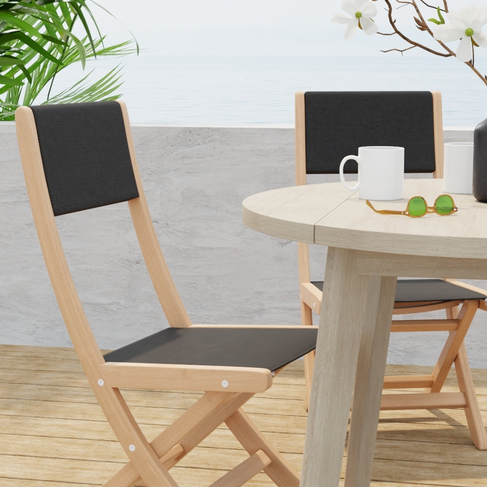Outdoor light-wood round dining set with black tweed fabric cushions; mug and green sunglasses on the table, plants & sea behind.