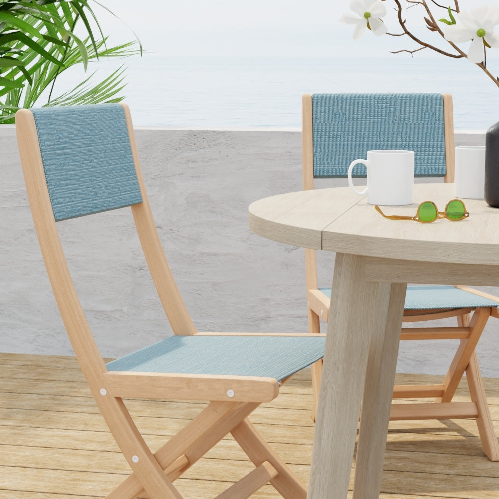 Light wood outdoor dining set with tweed cushions, round table and folding chairs on a wooden deck by the sea.
