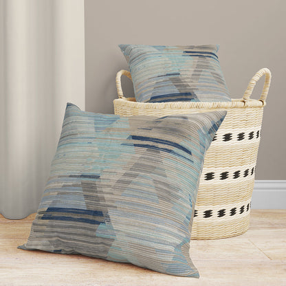 Two blue-gray abstract-pattern fabric-covered throw pillows with a textured weave beside a seagrass basket on a light wood floor.