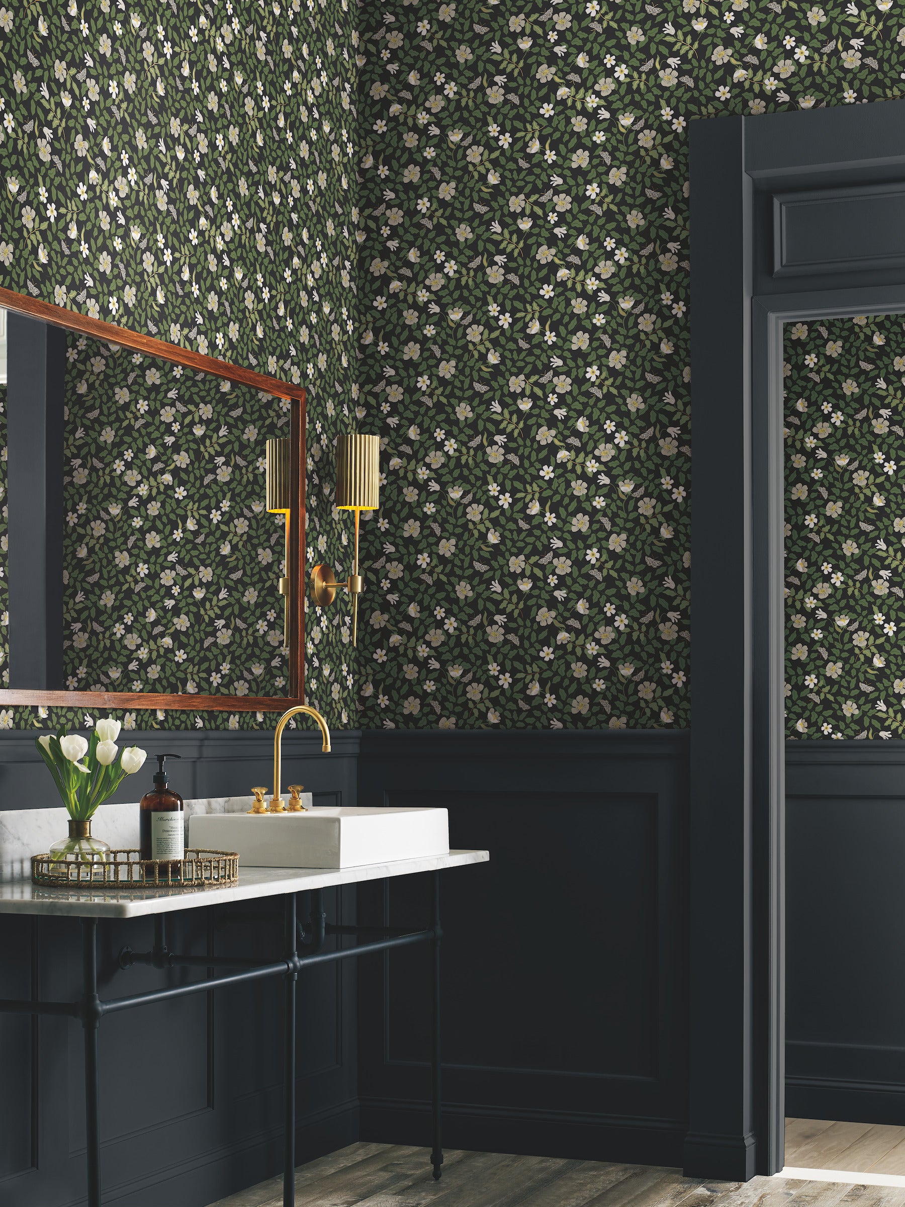 Floral wallpaper featuring primrose pattern in off-white & black tones, shown in a bathroom with a white sink, brass faucet, gold sconces, navy wainscoting, & a wooden mirror.