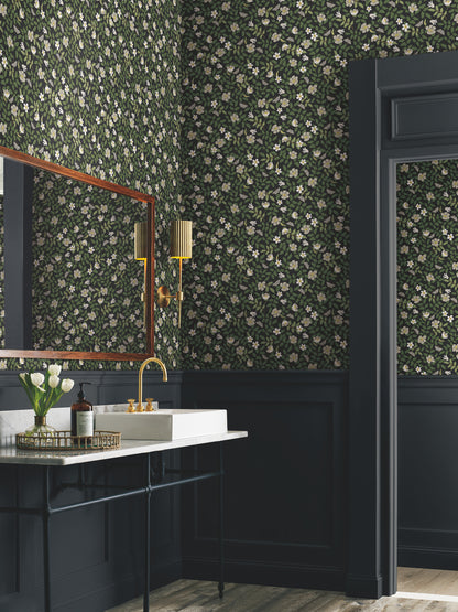 Floral wallpaper featuring primrose pattern in off-white & black tones, shown in a bathroom with a white sink, brass faucet, gold sconces, navy wainscoting, & a wooden mirror.