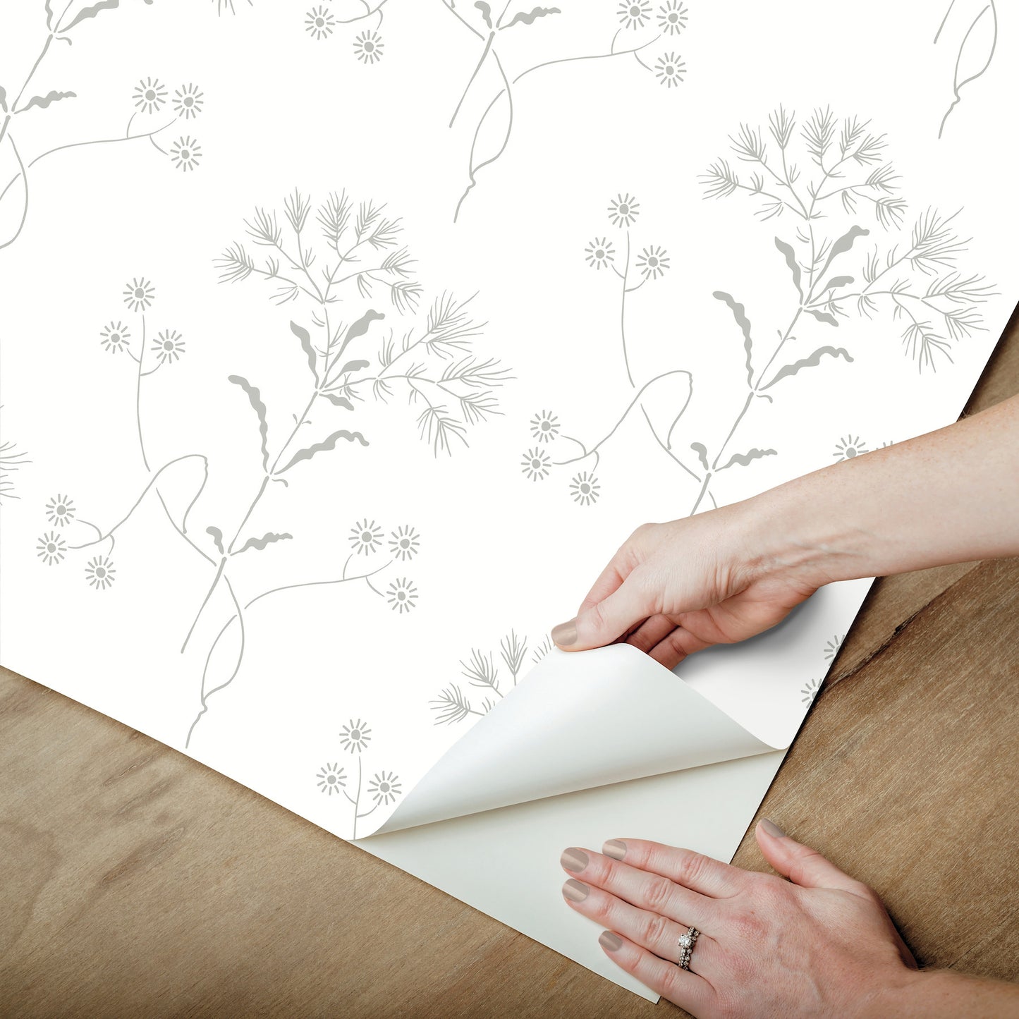 Gray wallpaper with botanical stems and tiny flowers; hands peel back a corner to reveal the backing.