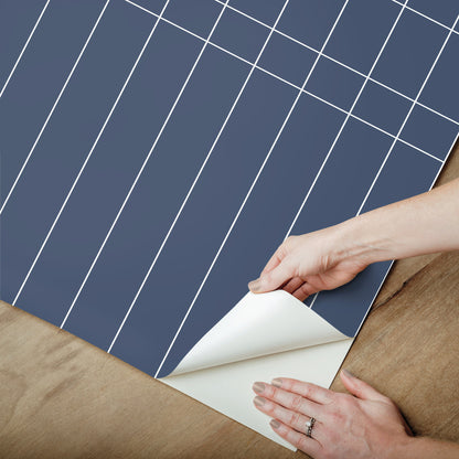 Blue grid-pattern wallpaper being peeled back to reveal white backing, hands at corner.