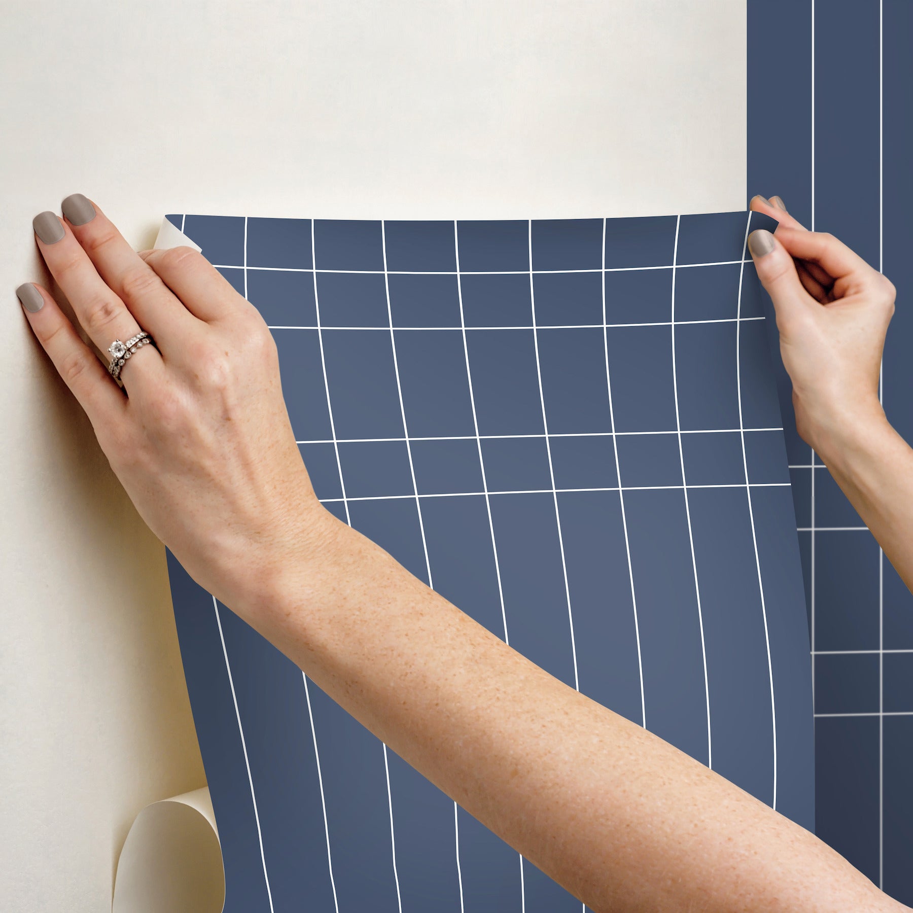 Two hands smooth a blue grid-pattern wallpaper sheet onto a wall.