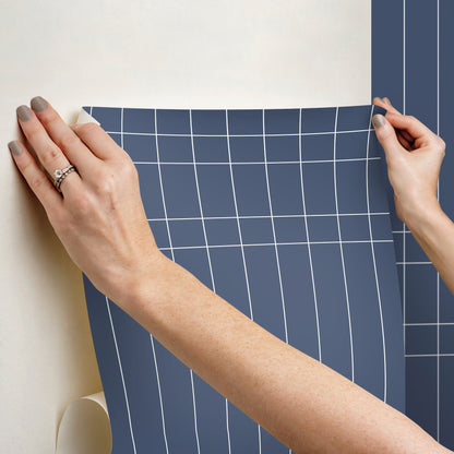 Two hands smooth a blue grid-pattern wallpaper sheet onto a wall.