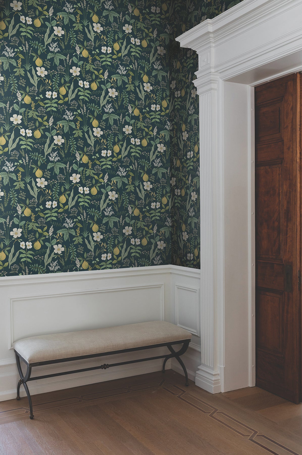 Room with navy botanical wallpaper, white wainscoting, beige cushioned bench with metal legs, and a dark wooden door.