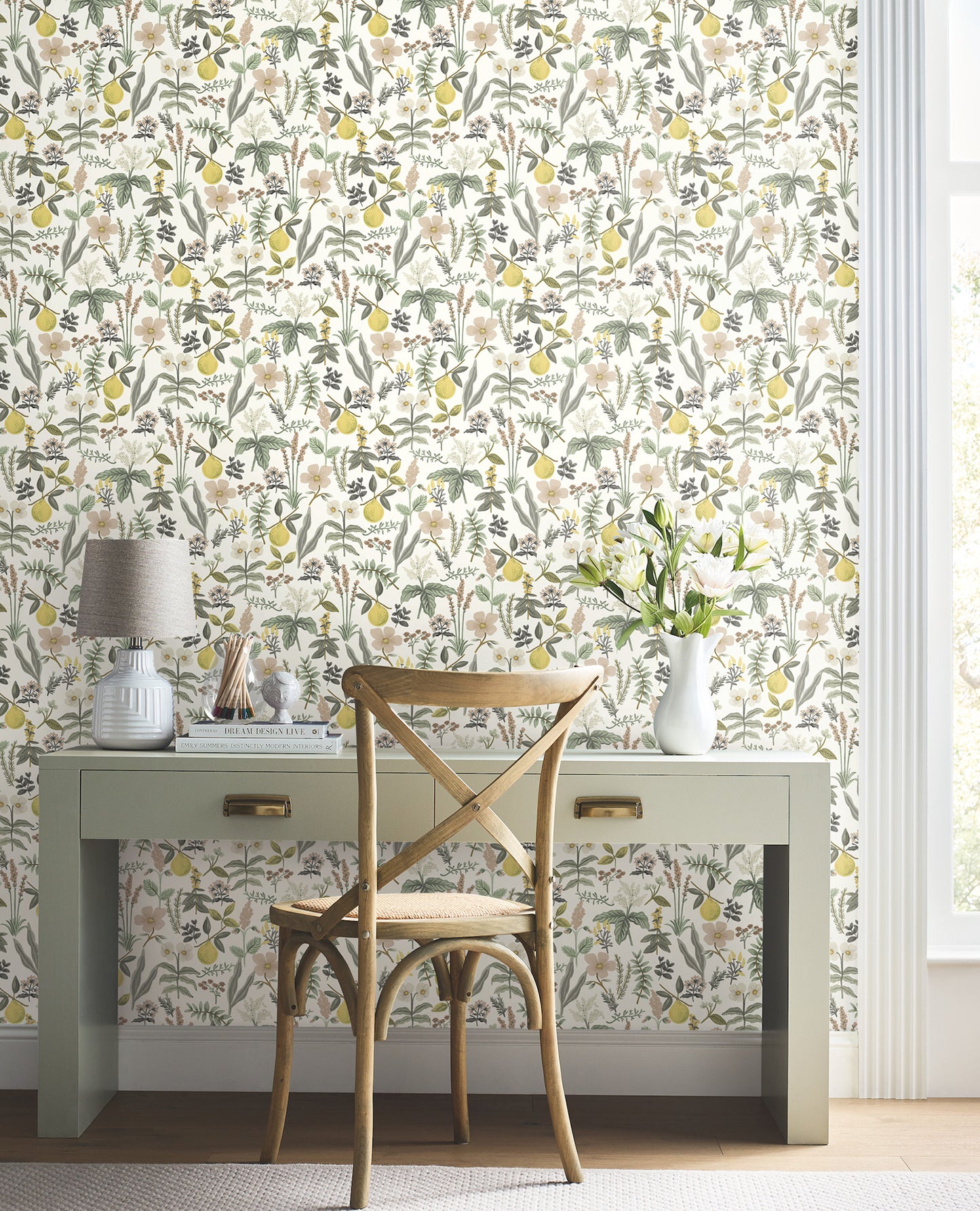 Light sage desk with a woven light wood cross-back chair against lemon-olive botanical wallpaper; lamp, books and white vase with flowers.