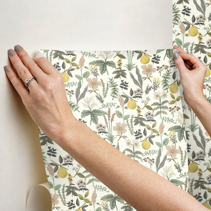 Hands smooth peel-and-stick floral wallpaper featuring yellow lemons, green leaves, and pale pink flowers onto a wall.