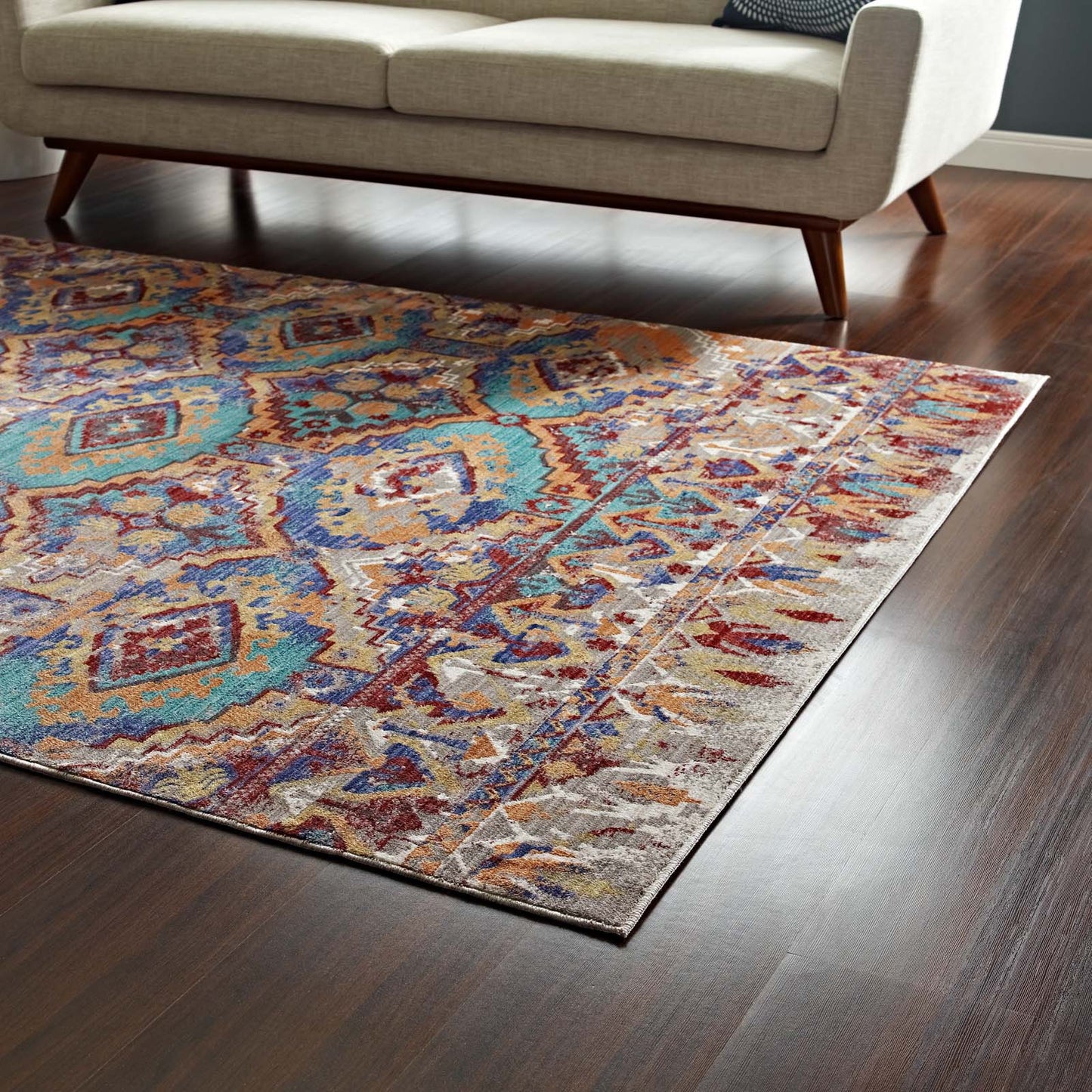Southwestern Aztec multicolored area rug in teal, blue, and orange with a beige border beneath a light sofa on a dark wood floor.