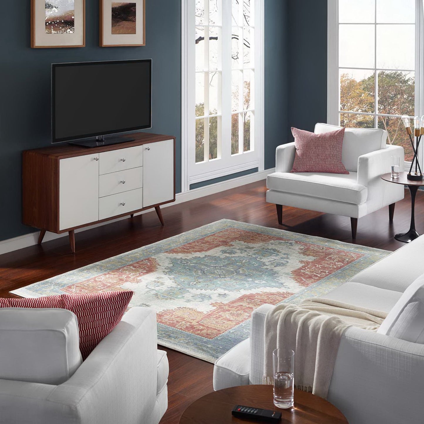 Cozy mid-century living room: blue walls, white sofa, distressed Southwestern Aztec multicolored area rug, wood-and-white TV console, framed art, windows.