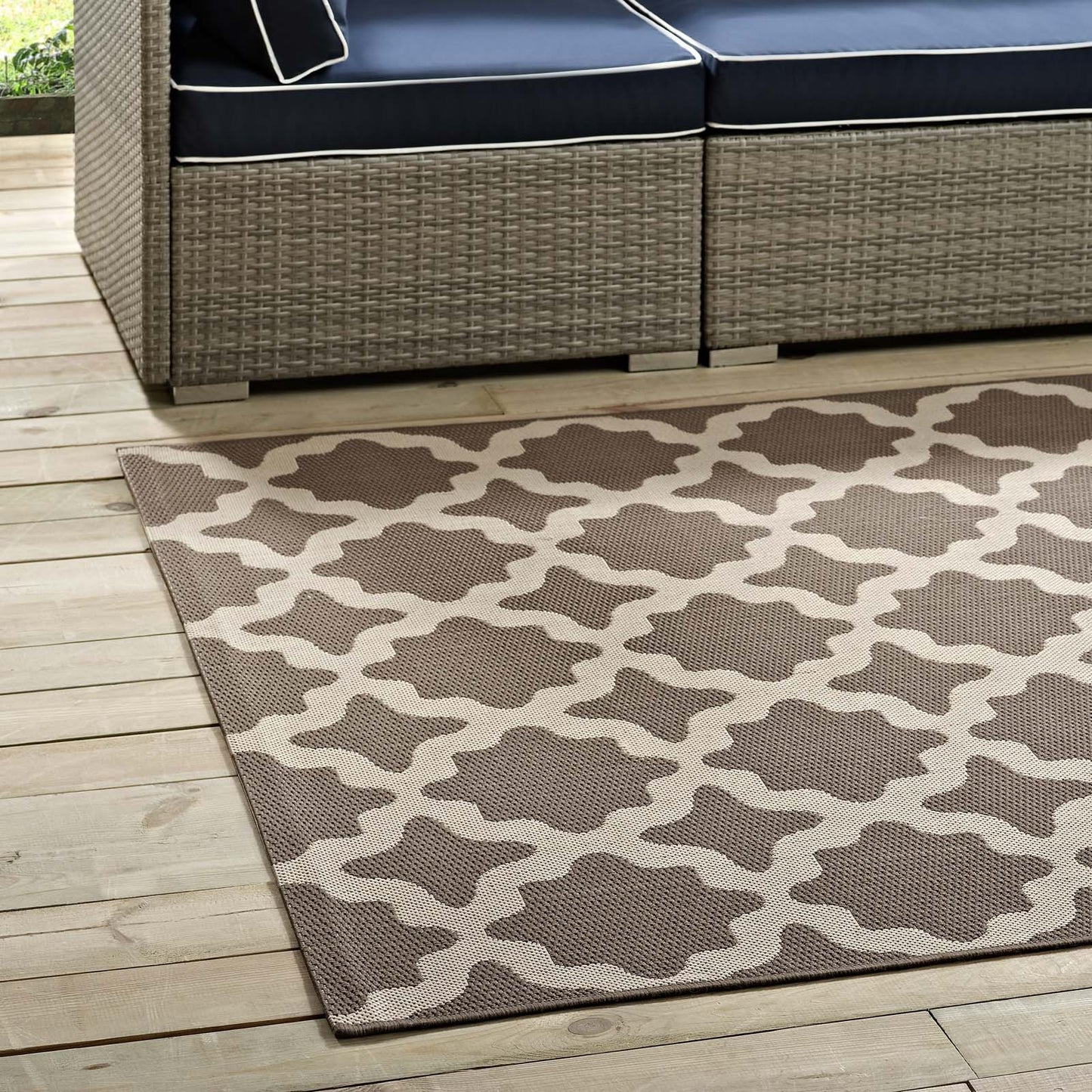Outdoor woven rug with beige lattice pattern on brown, laid on a wooden deck beside navy-cushioned wicker seating.