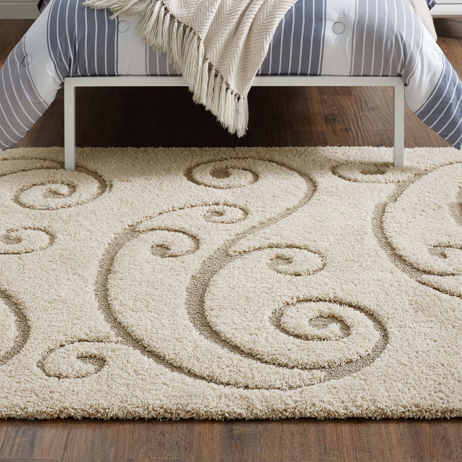 Beige shag rug with curling spiral motifs under a low bed with blue striped bedding on dark wood floor.