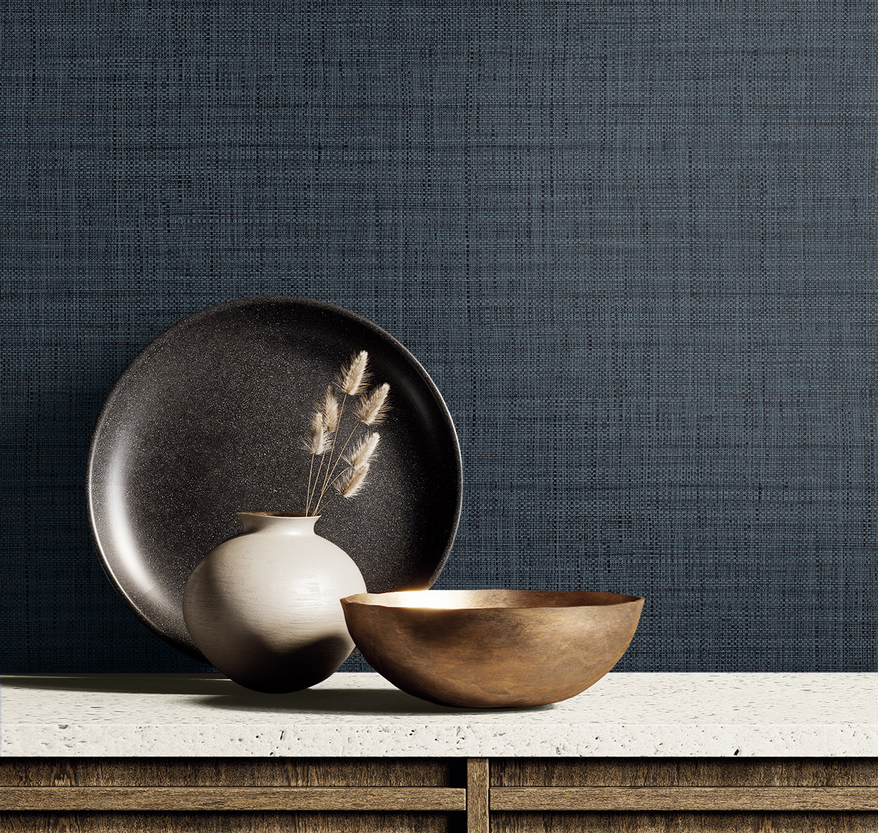 Dark blue textured wall with a beige ceramic vase and dried grasses, a black plate and a bronze bowl on a light countertop.
