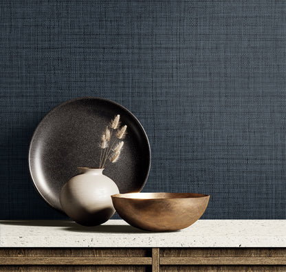 Dark blue textured wall with a beige ceramic vase and dried grasses, a black plate and a bronze bowl on a light countertop.