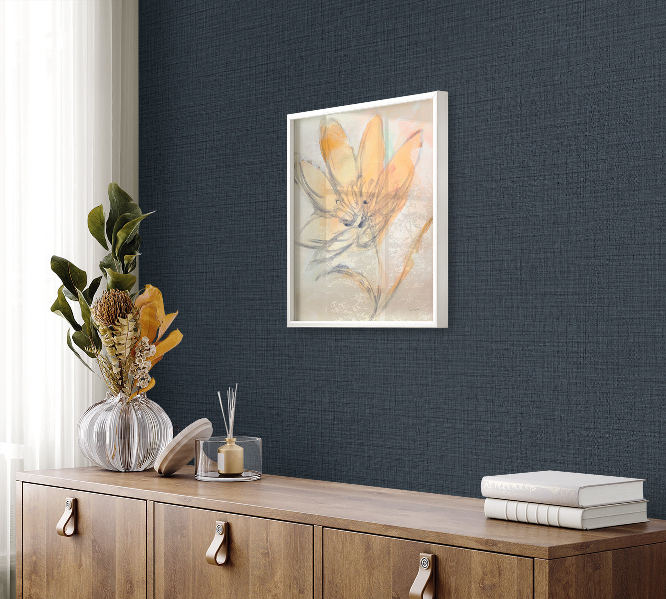Framed botanical print with orange petals on pale background, white frame, navy wall above sideboard with vase and books.