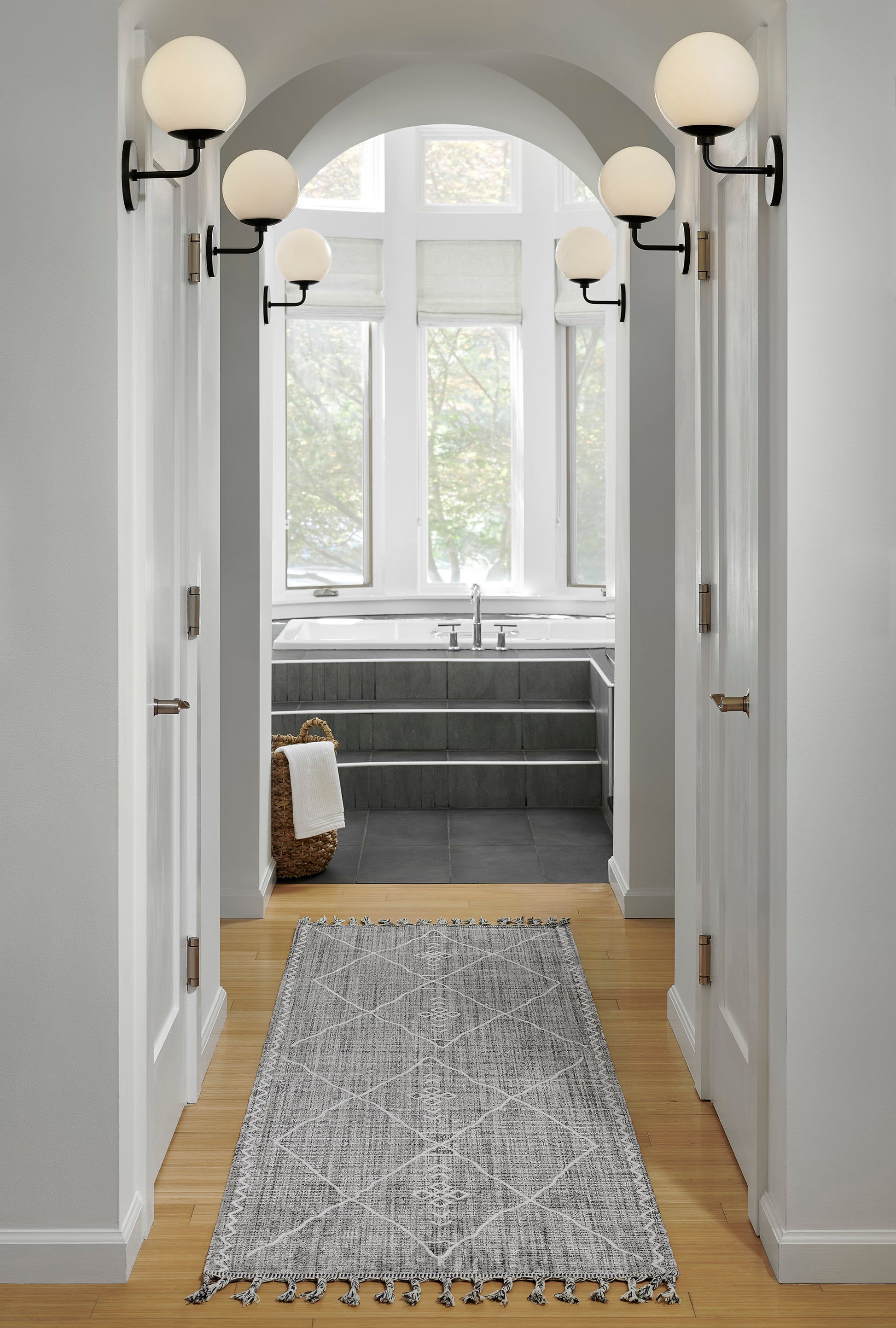 Gray geometric tassel rug runner in a white hallway to a bathroom with a dark tub, arched window, and globe sconces.