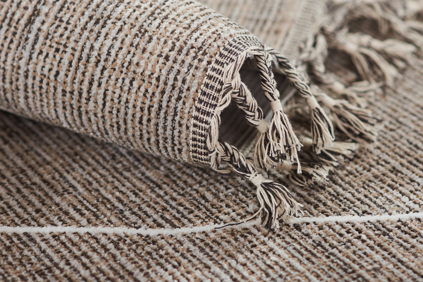 Beige and gray striped woven rug with tassel fringe along the edge, rolled at the corner.