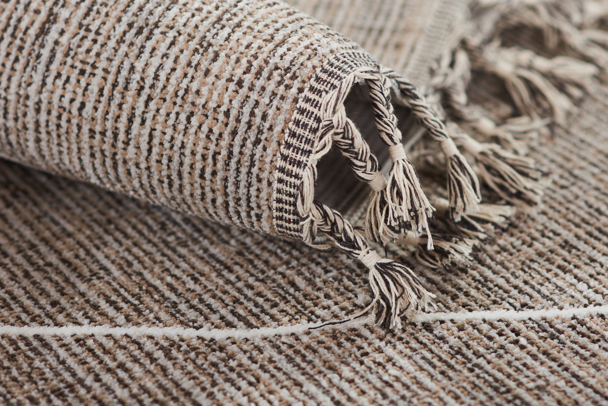 Beige and gray striped woven rug with tassel fringe along the edge, rolled at the corner.