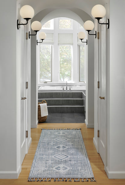 Hallway with white trim, globe sconces, light blue geometric runner on wooden floor, leads to bath with arched window.