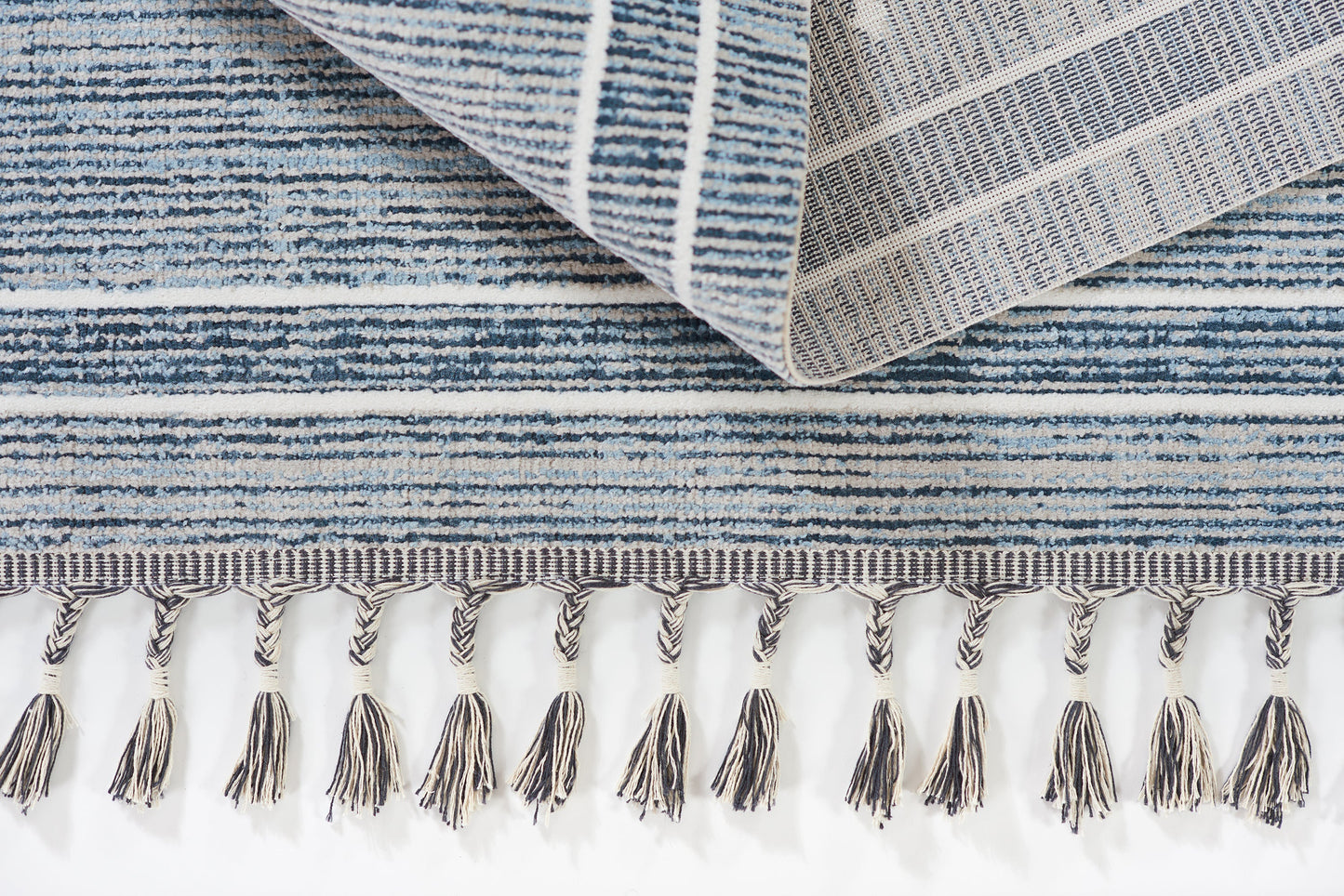 Blue striped woven rug with beige tassel fringe along edge; folded corner reveals denim tones.