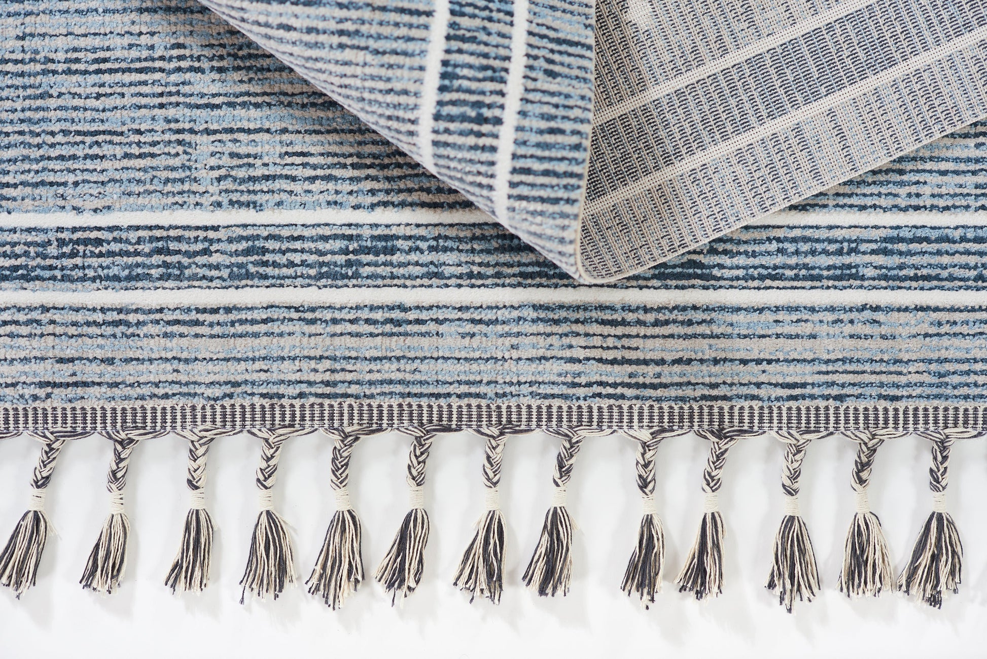 Blue striped woven rug with beige tassel fringe along edge; folded corner reveals denim tones.