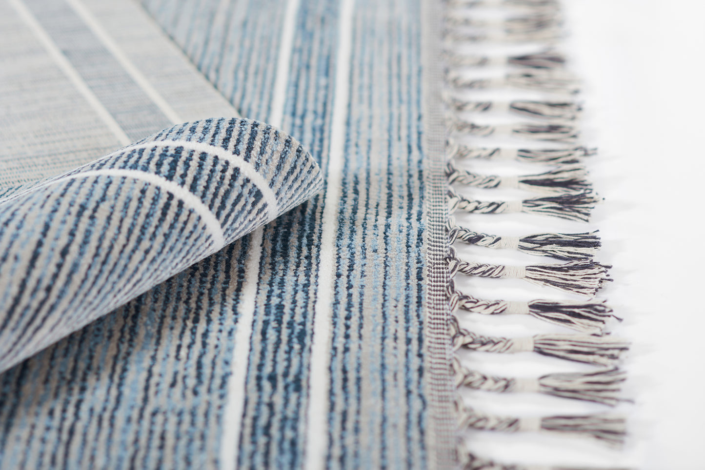 Blue striped woven rug with white fringe along edge, folded corner reveals soft textured weave.