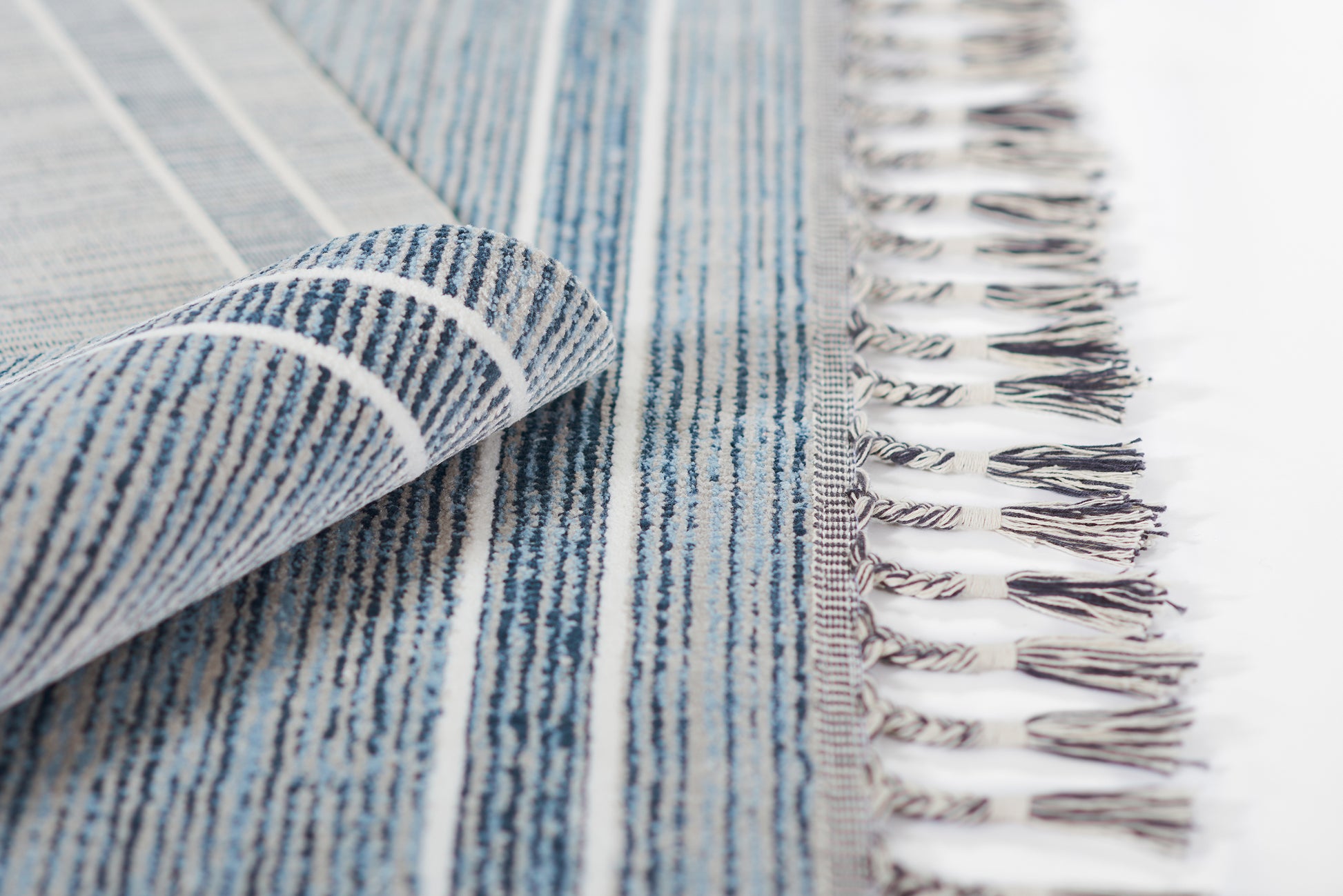 Blue striped woven rug with white fringe along edge, folded corner reveals soft textured weave.