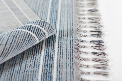 Blue striped woven rug with white fringe along edge, folded corner reveals soft textured weave.