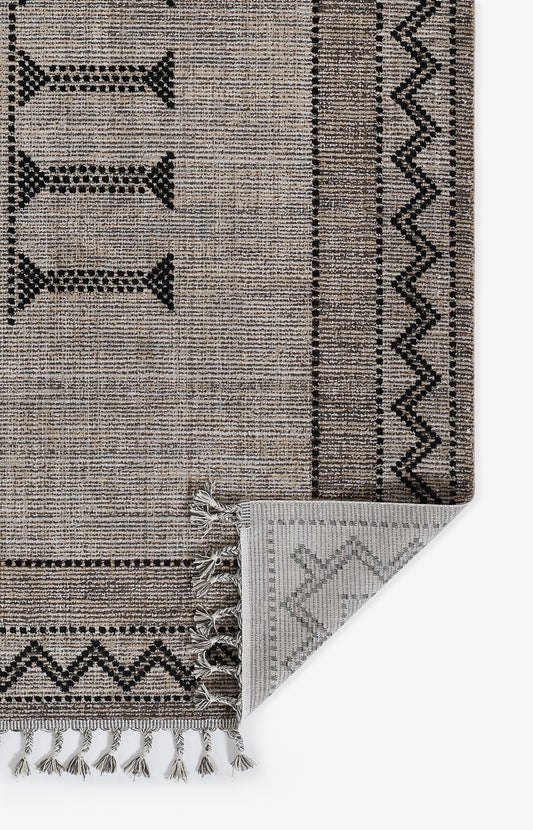 Beige-brown woven rug with black geometric borders, zigzag trim & tassel fringe; folded corner reveals gray underside.