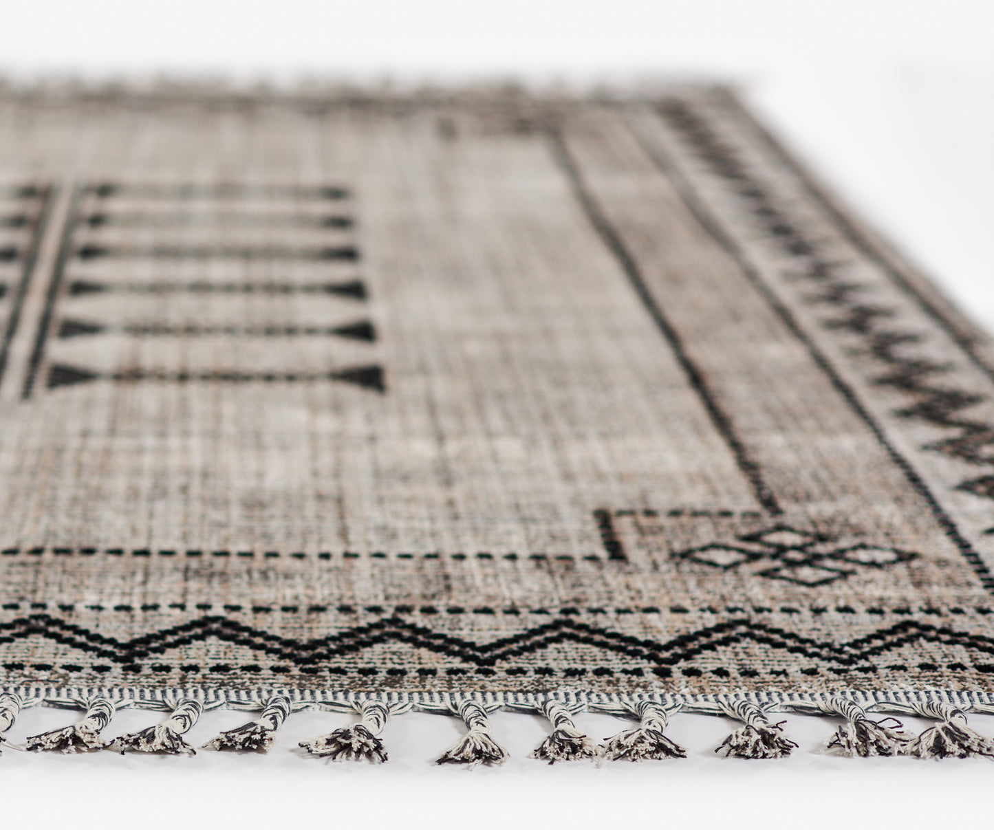 Beige-brown woven rug with a black geometric tribal pattern and fringed tassels along the edge.