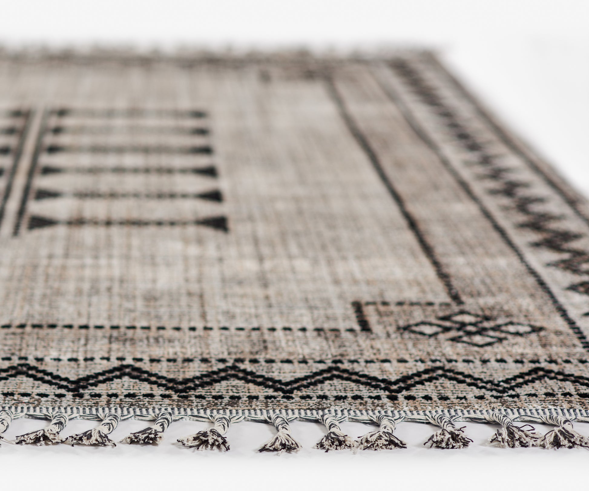 Beige-brown woven rug with a black geometric tribal pattern and fringed tassels along the edge.