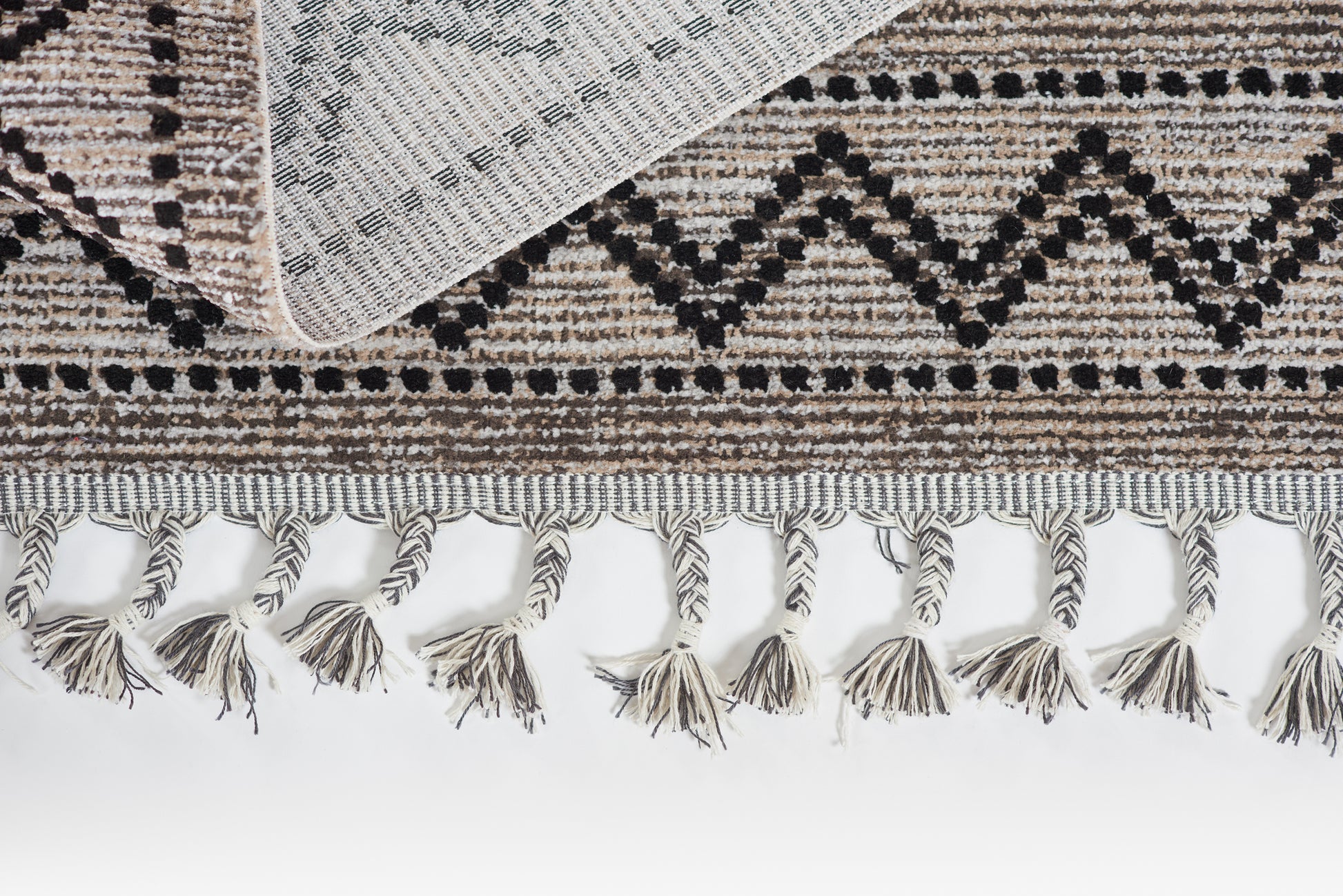 Beige and black geometric flat-weave rug with tassel fringe along edge; folded corner reveals underside.
