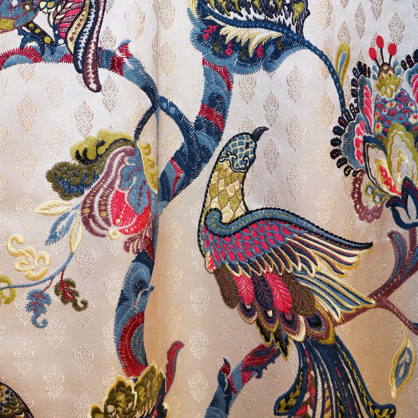 Ornate embroidered bird motif on cream damask fabric; vivid blues, pinks, yellows with floral vines.