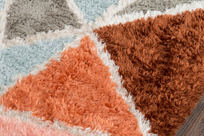 Shag area rug featuring bold geometric triangles in coral orange, peach, dusty blue, light gray, and brown for a retro look.