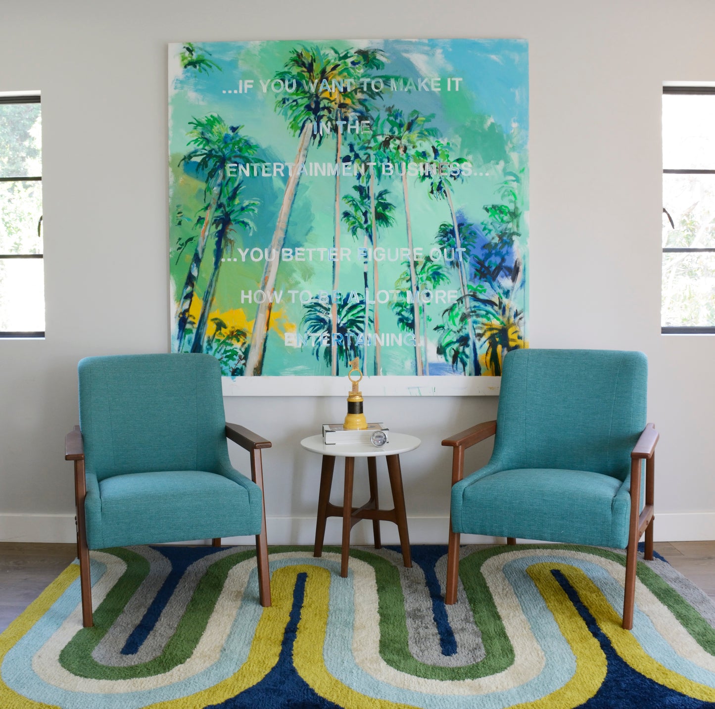 Two teal mid-century armchairs with wooden legs flank a round white side table under tropical palm art on a colorful rug.