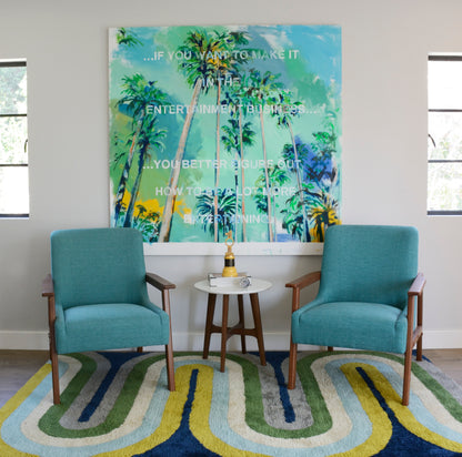 Two teal mid-century armchairs with wooden legs flank a round white side table under tropical palm art on a colorful rug.