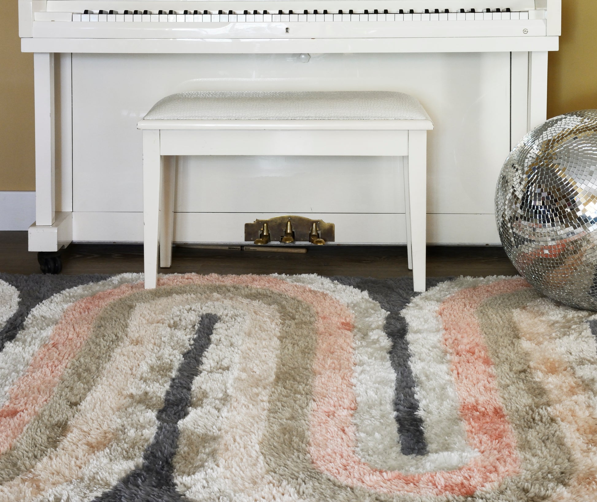 White upright piano with matching bench, brass pedals, on a plush multicolor shag rug beside a disco ball.